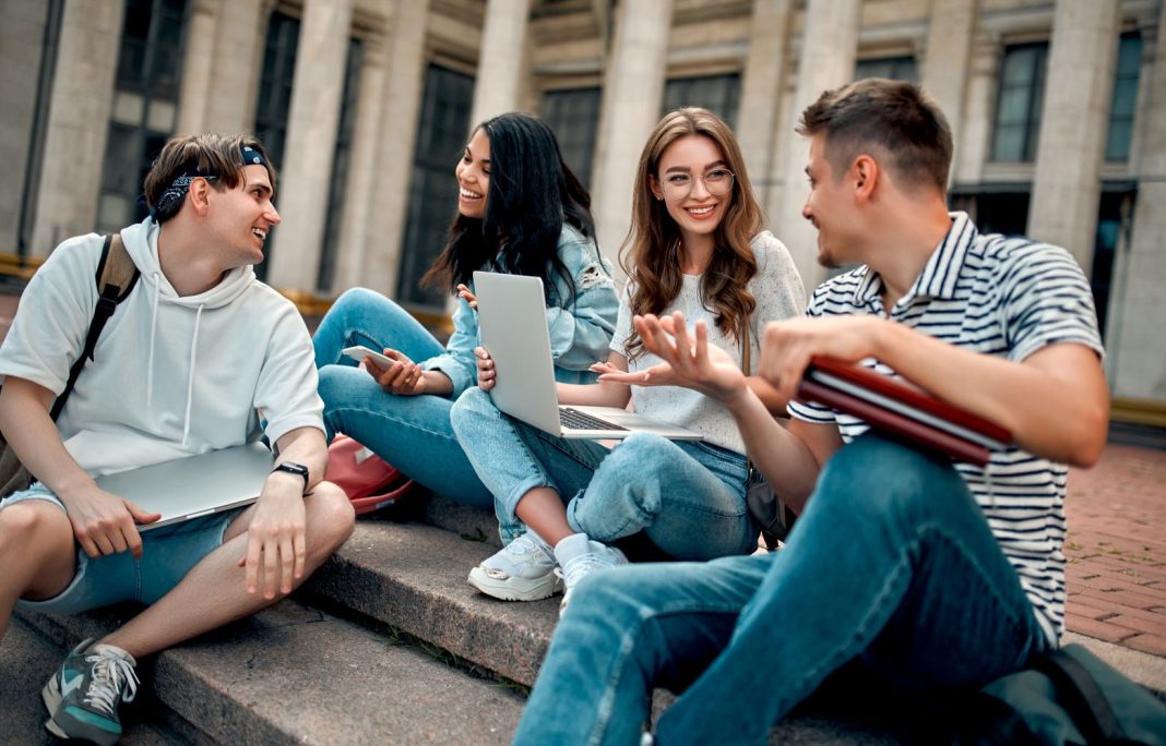 Group of college students sitting on campus steps with laptops and books, talking and studying together in front of a university building.