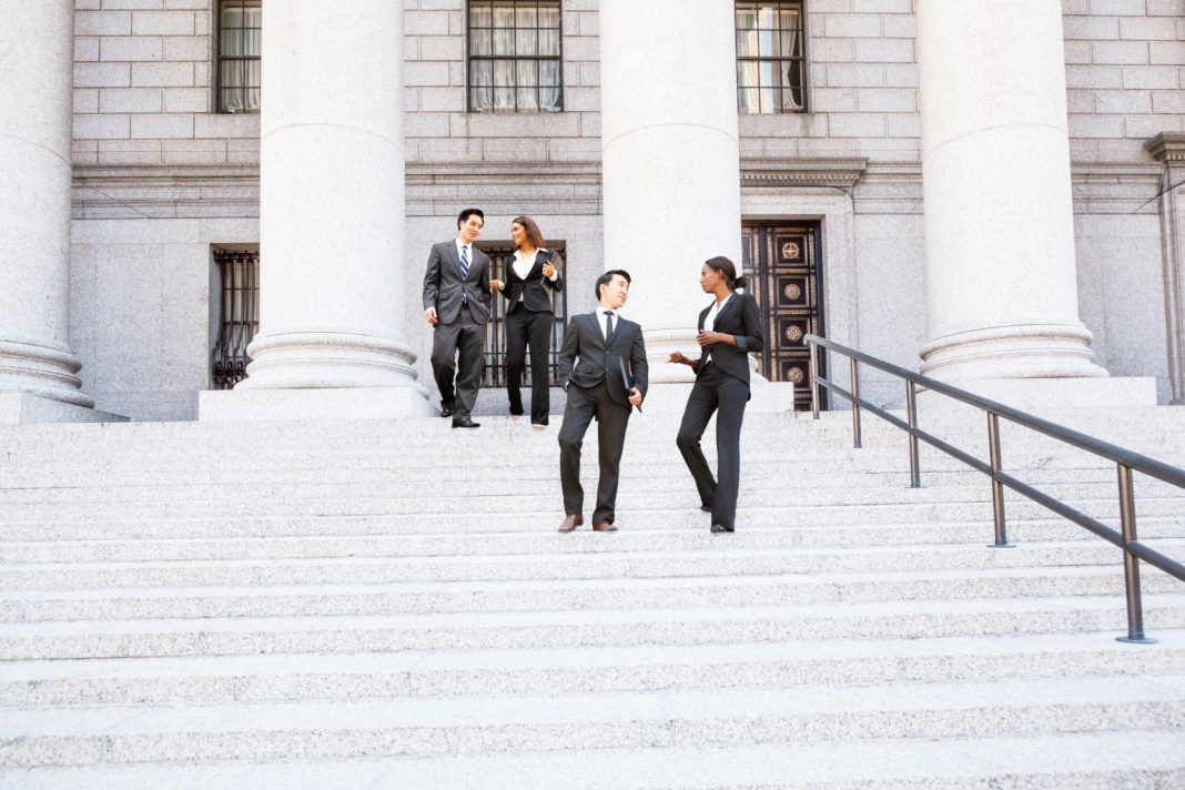 Four young professionals in business attire walk down the stone steps of a courthouse with tall columns while talking together. The group carries folders and appears to be leaving a legal proceeding or meeting.