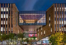 Modern law school building with tall stone walls and a glass skybridge connecting two sections above an open walkway. The illuminated structure stands at dusk with trees and pathways in the courtyard below.